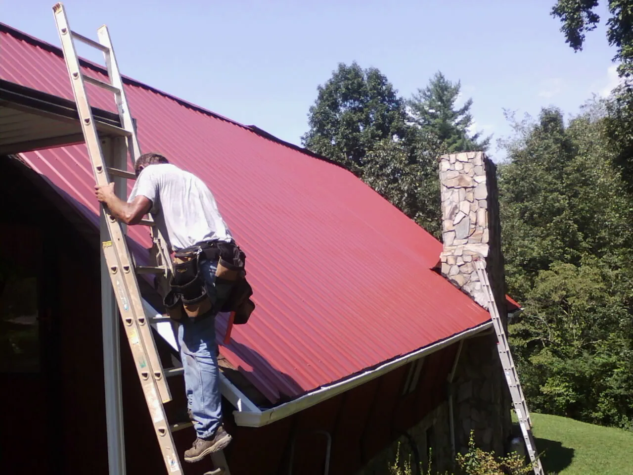 Roofer on a ladder beside a red metal roof and stone chimney — Soffit & Fascia Repair by Roofing Brothers in Mertztown